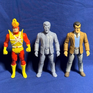 May include: Three action figures are displayed against a blue backdrop. The figures include a character with a flaming head and red and yellow suit, a gray suited figure, and a figure in a brown jacket and glasses. The figures are approximately 6 inches tall.