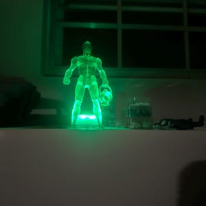 May include: A translucent green action figure of a superhero, illuminated with green light. The figure is holding a lantern and standing on a small platform. The background is dark, with a window visible.