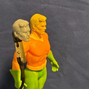May include: A detailed action figure of a superhero, featuring an orange top, green pants, and green gloves. The figure has blonde hair and a determined expression. A gray head is held by a clamp next to the figure.