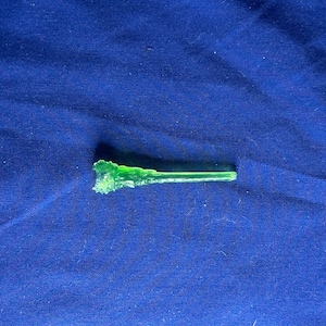 May include: A small, translucent, neon green glass wand with a textured, irregular tip. The wand is lying on a dark blue fabric surface. The wand's design is simple and elegant.