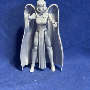 Puede incluir: Figura de acción gris de un personaje femenino con capucha y capa. La figura está en una pose dinámica con los brazos extendidos. La figura está de pie sobre una superficie azul.