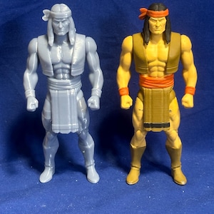 May include: Two action figures of a Native American warrior. One is gray, the other is tan with a red headband and accents. Both figures have long hair, headbands, and traditional attire. The figures are standing on a blue surface.