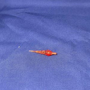 Könnte beinhalten: Ein kleines, rot-oranges Dekorationsobjekt mit spitzem Ende und bauchigem Zentrum. Das Objekt befindet sich vor einem einfarbigen blauen Hintergrund, wodurch ein Farbkontrast entsteht. Der Artikel scheint aus Glas oder einem ähnlichen Material zu bestehen.