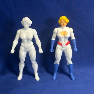 Pode incluir: Duas figuras de ação de uma super-heroína. Uma é toda branca, a outra é branca com detalhes em azul, vermelho e amarelo. Ambas as figuras estão em uma pose dinâmica, com escultura detalhada e uma estética clássica de super-herói.
