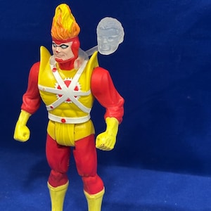 May include: A Flamebird action figure with a yellow and red costume. The figure has a flame-shaped headpiece, yellow gloves and boots, and a clear plastic head attachment. The figure is posed against a blue background.