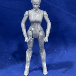 Puede incluir: Figura de acción articulada gris de un personaje femenino, de pie con los puños cerrados. La figura tiene un corte de pelo corto y lleva un atuendo ajustado con elementos detallados similares a una armadura. Sobre un fondo azul.