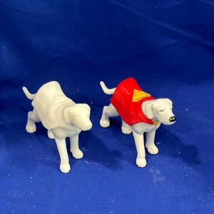 Puede incluir: Dos figuras de perros blancos. Uno es liso, el otro lleva una capa roja con un emblema amarillo. Ambos perros est&aacute;n de pie sobre una superficie azul. Las figuras parecen ser de pl&aacute;stico.
