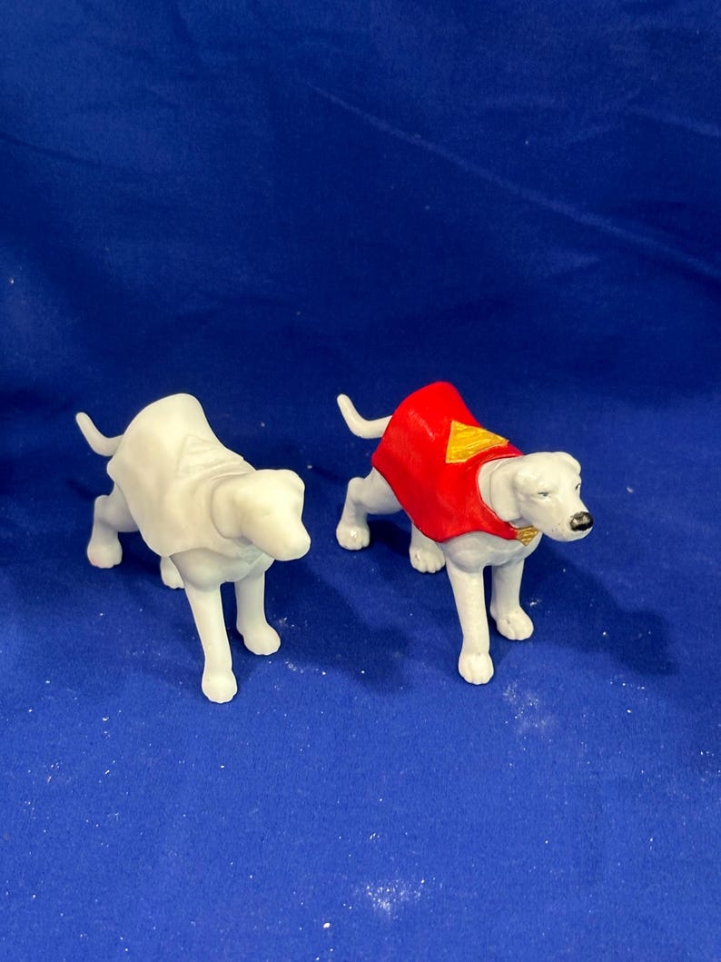 Puede incluir: Dos figuras de perros blancos. Uno es liso, el otro lleva una capa roja con un emblema amarillo. Ambos perros est&aacute;n de pie sobre una superficie azul. Las figuras parecen ser de pl&aacute;stico.