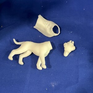 Puede incluir: Una figura de perro desmontada, de color blanquecino, con piezas separadas para la cabeza, el cuerpo y las patas. El perro est&aacute; en posici&oacute;n de caminar. El fondo es una superficie azul lisa.