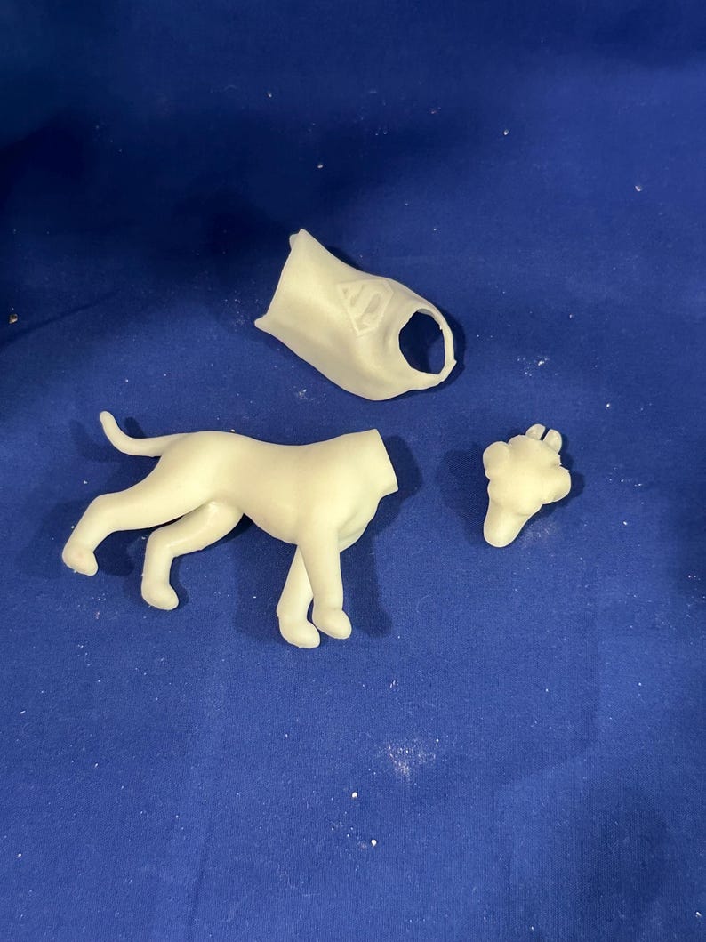 Puede incluir: Una figura de perro desmontada, de color blanquecino, con piezas separadas para la cabeza, el cuerpo y las patas. El perro est&aacute; en posici&oacute;n de caminar. El fondo es una superficie azul lisa.