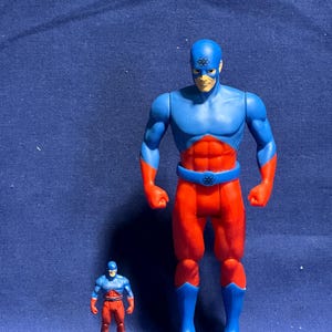 Pode incluir: Duas figuras de a&ccedil;&atilde;o de um super-her&oacute;i. A figura maior &eacute; azul e vermelha com um cinto e botas azuis. A figura menor tem o mesmo esquema de cores. Ambas as figuras t&ecirc;m um s&iacute;mbolo na testa.
