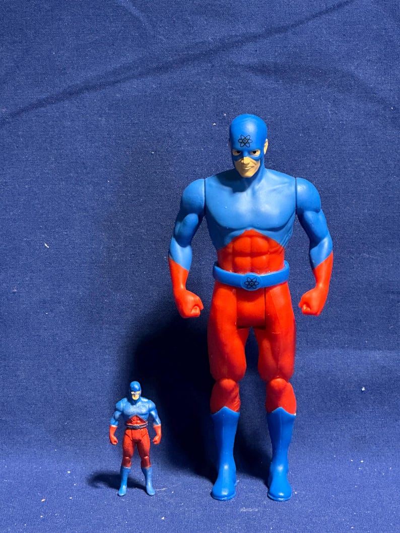 Pode incluir: Duas figuras de a&ccedil;&atilde;o de um super-her&oacute;i. A figura maior &eacute; azul e vermelha com um cinto e botas azuis. A figura menor tem o mesmo esquema de cores. Ambas as figuras t&ecirc;m um s&iacute;mbolo na testa.