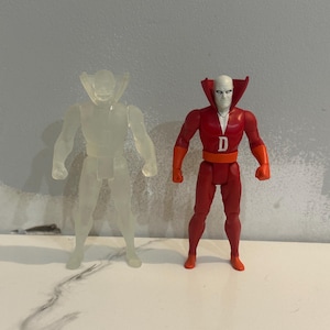 Peut inclure: Deux figurines d'action. L'une est translucide, l'autre est rouge avec des accents orange et un visage blanc. La figurine rouge porte un "D" blanc sur la poitrine. Les deux figurines sont sur une surface blanche.
