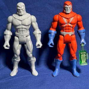 Puede incluir: Dos figuras de acción de personajes masculinos musculosos. Una es gris, la otra roja con detalles azules. La figura roja sostiene un objeto verde rectangular. Ambas figuras tienen expresiones serias y están de pie.