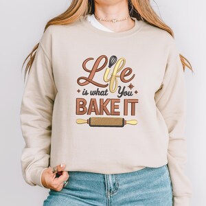 Pode incluir: Camisola bege com o texto "Life is what You BAKE IT" em letras castanhas e amarelas. O design inclui um batedor e um rolo de massa. A camisola &eacute; de cor neutra.