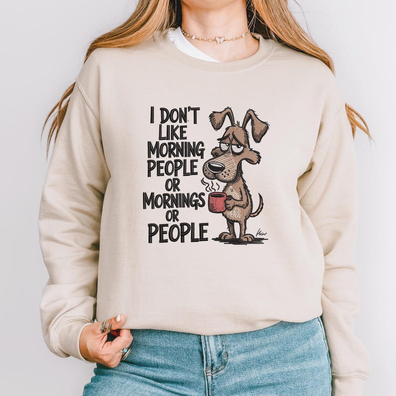 May include: Beige sweatshirt with a cartoon dog holding a coffee mug. The dog has a sleepy expression. Text on the sweatshirt reads "I don't like morning people or mornings or people."