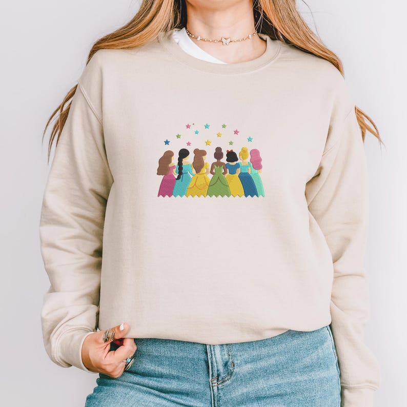 May include: Beige sweatshirt featuring a colorful illustration of seven cartoon princesses standing together under a rainbow of stars. The princesses are wearing gowns in various colors, including pink, blue, yellow, and green. The sweatshirt has a crew neck and long sleeves.