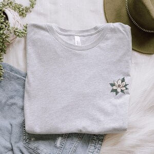 Pode incluir: Camiseta cinza claro com gola redonda, com um pequeno desenho de flor de magn&oacute;lia na frente. A magn&oacute;lia tem p&eacute;talas brancas e folhas verdes. A camiseta est&aacute; dobrada sobre uma superf&iacute;cie branca, com um chap&eacute;u verde e jeans.
