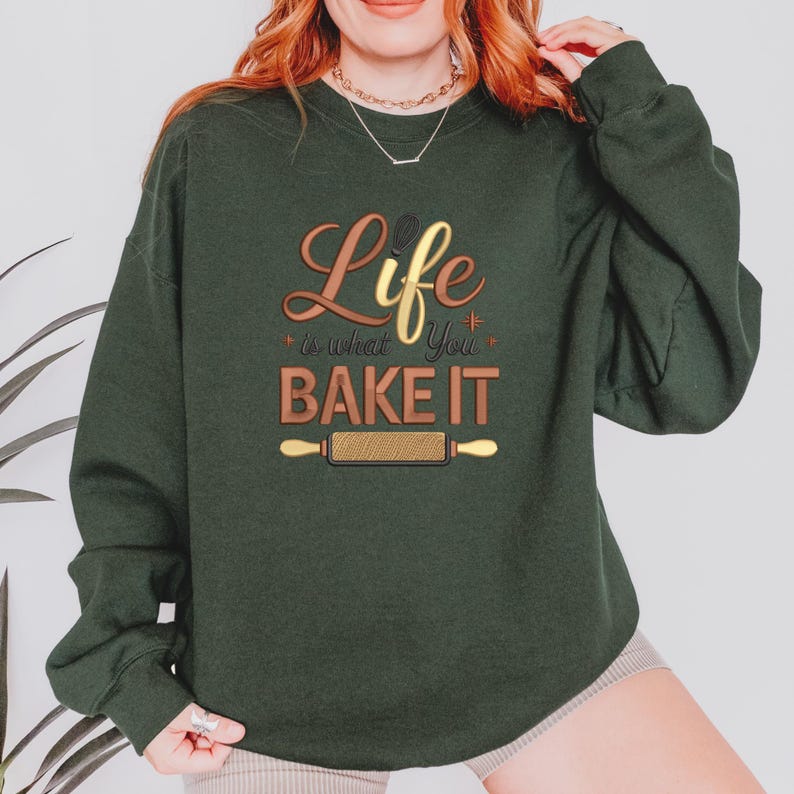 Pode incluir: Camisola verde escuro com o texto "Life is what you BAKE IT" em letras douradas e castanhas. Um gr&aacute;fico de rolo de massa est&aacute; abaixo do texto. A camisola &eacute; um estilo confort&aacute;vel e casual.