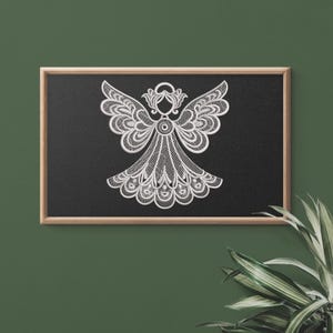May include: A white, detailed angel design on a black background, framed in wood. The angel features intricate wing and dress patterns, creating a delicate, lace-like appearance. The artwork is displayed against a dark green wall with a plant in the corner.
