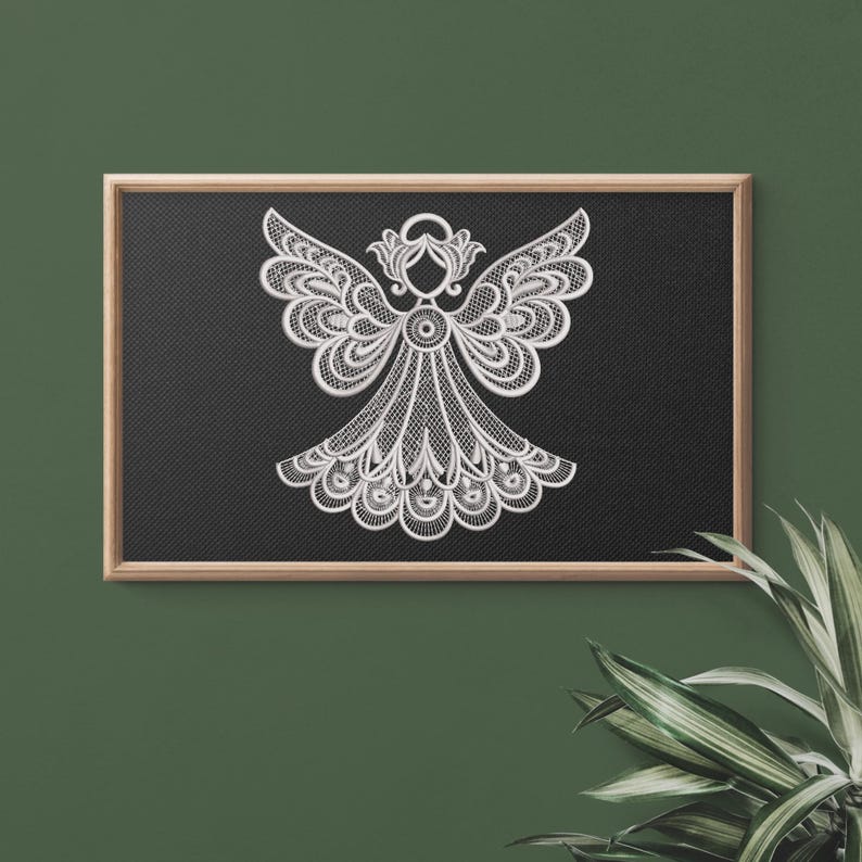 May include: A white, detailed angel design on a black background, framed in wood. The angel features intricate wing and dress patterns, creating a delicate, lace-like appearance. The artwork is displayed against a dark green wall with a plant in the corner.