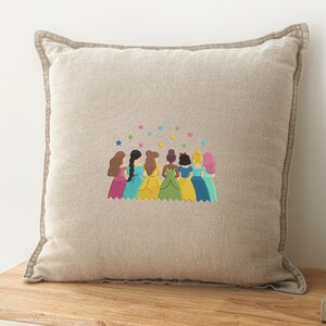 May include: Beige square throw pillow with embroidered design. The design features eight figures in colorful gowns with various hairstyles, under a scattering of colorful stars. The pillow has a light brown border.