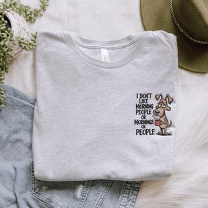 May include: Light gray t-shirt with the text "I don't like morning people or mornings or people." A cartoon dog holding a coffee cup is printed on the right side. The shirt is made of soft material.