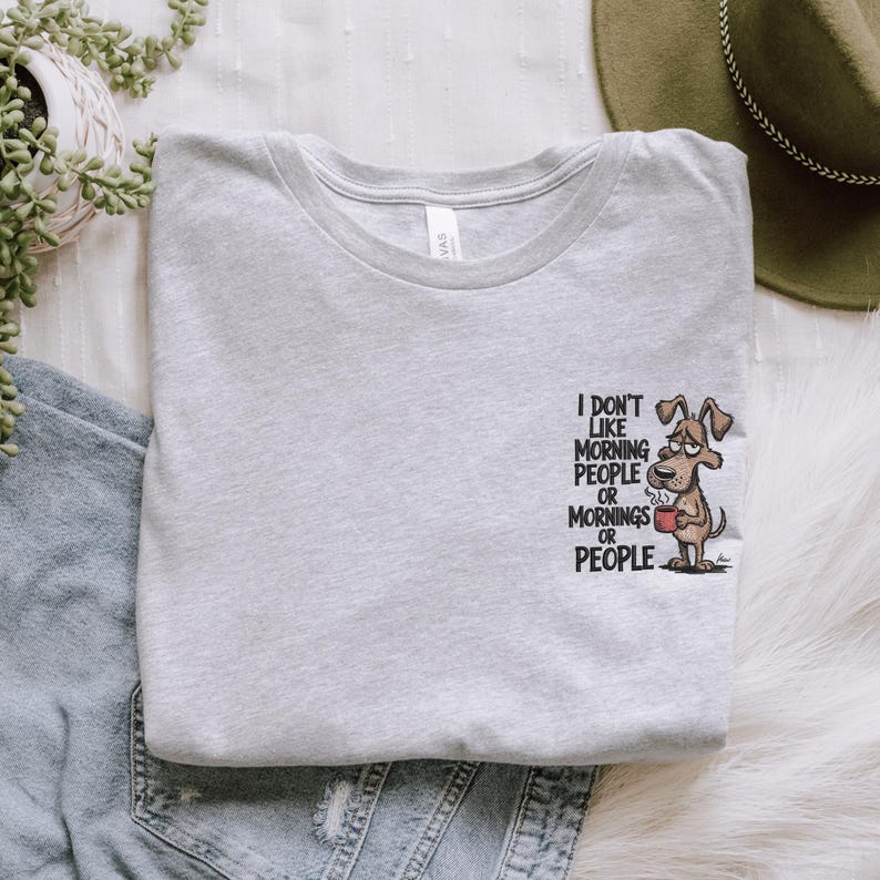 May include: Light gray t-shirt with the text "I don't like morning people or mornings or people." A cartoon dog holding a coffee cup is printed on the right side. The shirt is made of soft material.