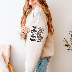 May include: Beige sweatshirt with the text "I don't like morning people or mornings or people" and a cartoon dog holding a mug. The dog has a grumpy expression. The sweatshirt is a neutral color.
