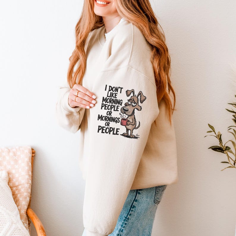 May include: Beige sweatshirt with the text "I don't like morning people or mornings or people" and a cartoon dog holding a mug. The dog has a grumpy expression. The sweatshirt is a neutral color.
