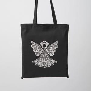 May include: Black tote bag featuring a white, detailed angel design. The angel has outstretched wings and a flowing gown, all rendered in a delicate, lace-like pattern. The bag has two black handles for carrying.