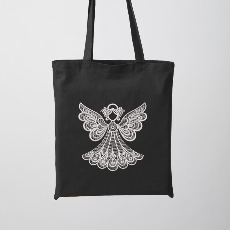 May include: Black tote bag featuring a white, detailed angel design. The angel has outstretched wings and a flowing gown, all rendered in a delicate, lace-like pattern. The bag has two black handles for carrying.