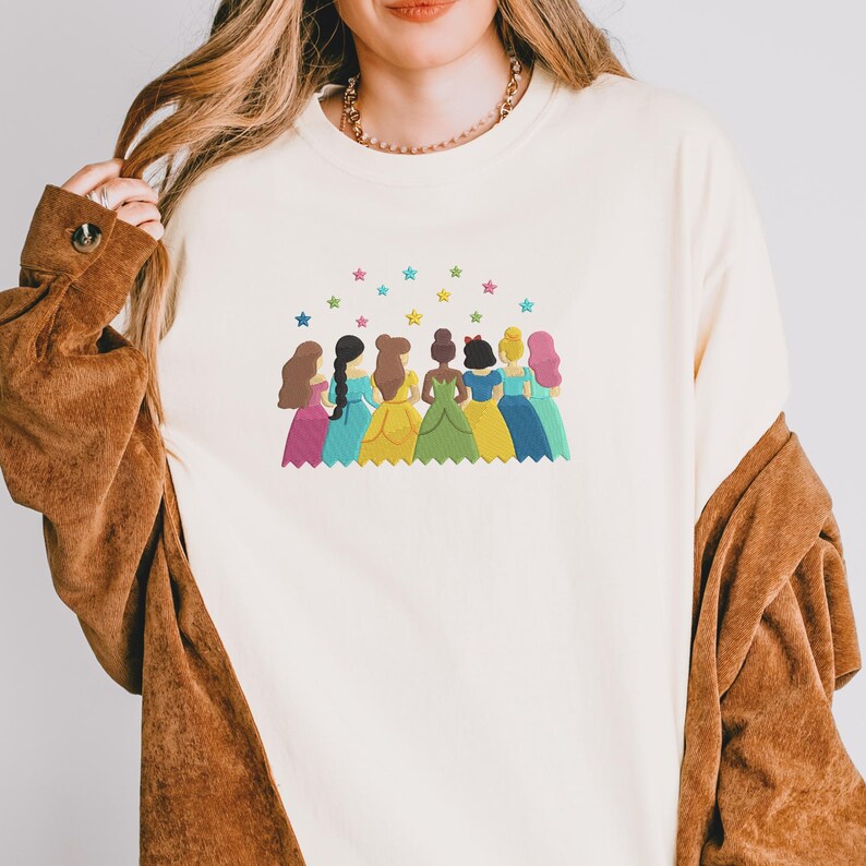 May include: Cream-colored sweatshirt featuring an embroidered design of seven cartoon princesses in colorful gowns. Above the princesses are colorful stars. The sweatshirt has long sleeves and a crew neck.