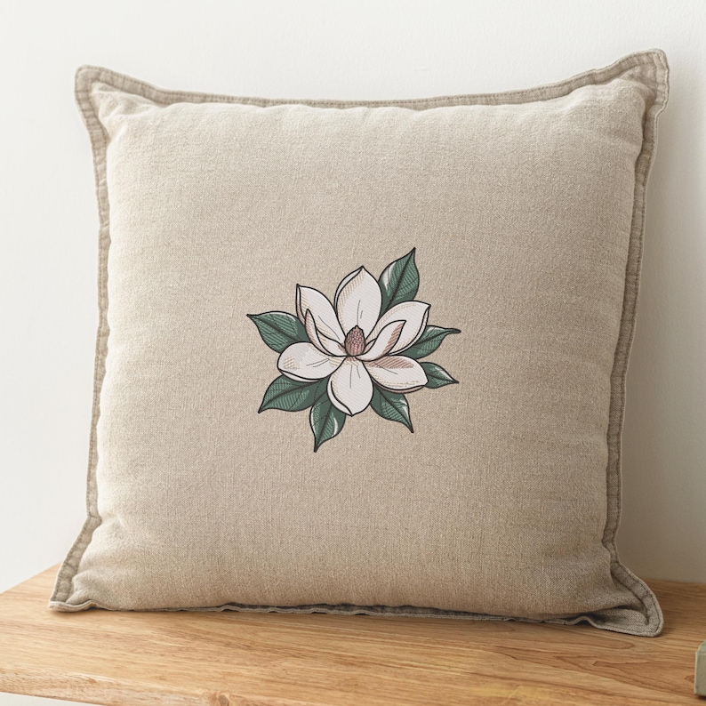 Pode incluir: Almofada quadrada bege com um bordado de flor de magn&oacute;lia. A flor tem p&eacute;talas brancas com um centro rosa, rodeadas por folhas verdes. A almofada tem uma borda castanha clara.