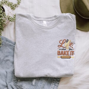Pode incluir: Uma camiseta cinza claro com a frase bordada "Life is what you BAKE IT" em marrom, dourado e laranja. O design inclui um rolo de massa e outros gr&aacute;ficos relacionados &agrave; panifica&ccedil;&atilde;o. A camiseta est&aacute; dobrada e exibida em uma superf&iacute;cie branca.