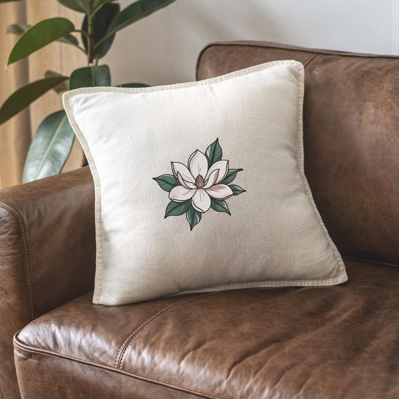 Pode incluir: Almofada quadrada com fundo creme e um desenho de flor de magn&oacute;lia. A flor tem p&eacute;talas brancas, centro rosa e folhas verdes. A almofada est&aacute; em um sof&aacute; de couro marrom.