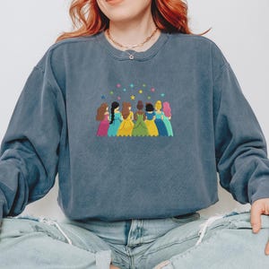 May include: A blue sweatshirt with an embroidered design of seven figures in colorful dresses, with stars above them. The figures have different skin tones and hair colors. The sweatshirt has a crew neck and long sleeves.