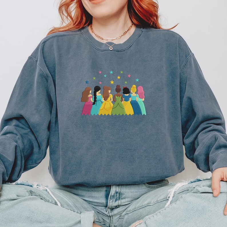 May include: A blue sweatshirt with an embroidered design of seven figures in colorful dresses, with stars above them. The figures have different skin tones and hair colors. The sweatshirt has a crew neck and long sleeves.