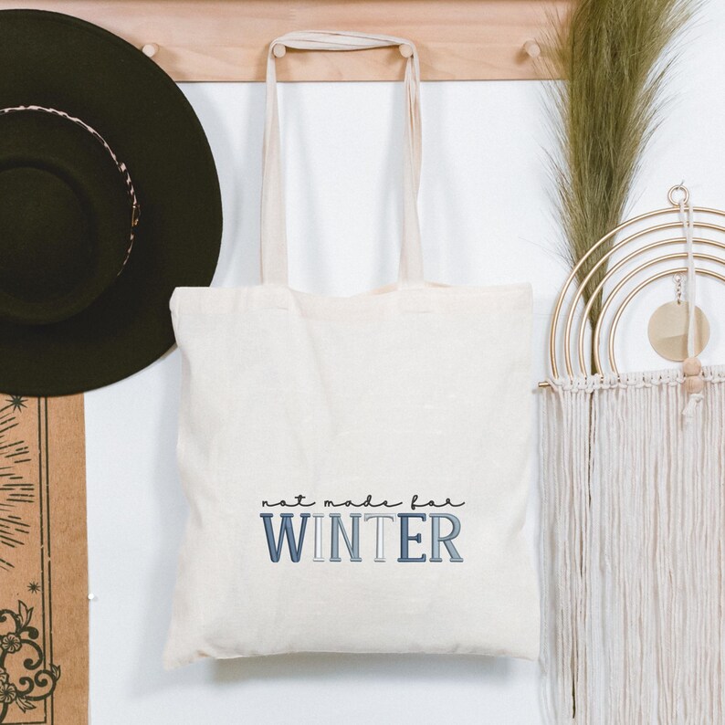 Puede incluir: Bolso tote de color crema con el texto "not made for WINTER" en letras azules y grises. El bolso cuelga de un perchero de madera, junto a un sombrero negro y adornos de pared decorativos. El bolso tiene asas largas de color crema.