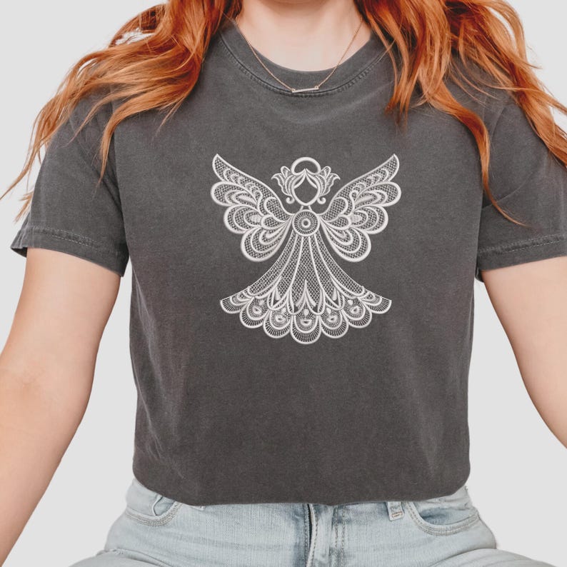 May include: Dark gray t-shirt featuring a detailed white angel design. The angel has outstretched wings and a flowing gown, creating a delicate and intricate pattern. The shirt is a comfortable, casual style.