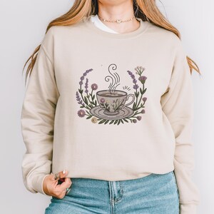 May include: Beige sweatshirt with an embroidered design of a teacup on a saucer, encircled by flowers and lavender. The teacup has steam rising from it. The design is in shades of purple, green, and beige.