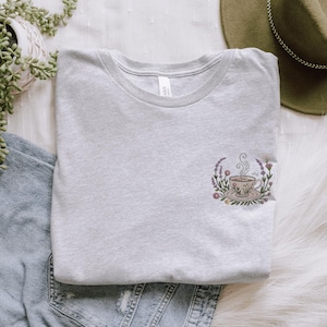 May include: A pale grey t-shirt featuring an embroidered teacup design with steam, encircled by flowers and lavender. The shirt is folded on a white surface, with jeans and a green hat partially visible. The design is approximately 5 cm in size.