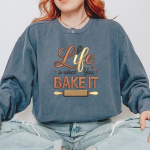 Pode incluir: Um moletom azul-acinzentado com o texto "Life is what You BAKE IT" em letras douradas e marrons. O design inclui um batedor e um rolo de massa. O moletom &eacute; um estilo casual e confort&aacute;vel.