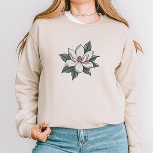 Pode incluir: Camisola bege com um design floral. O gr&aacute;fico &eacute; uma flor de magn&oacute;lia branca com folhas verdes e um centro rosa. A camisola &eacute; de cor neutra e tem gola redonda.