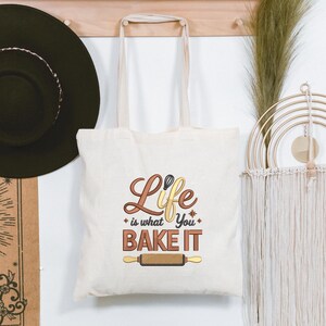 Pode incluir: Saco tote creme com o texto "Life is what you bake it" em letras castanhas e amarelas. O design inclui um batedor e um rolo de massa. O saco tem al&ccedil;as creme e est&aacute; pendurado num cabide de madeira.