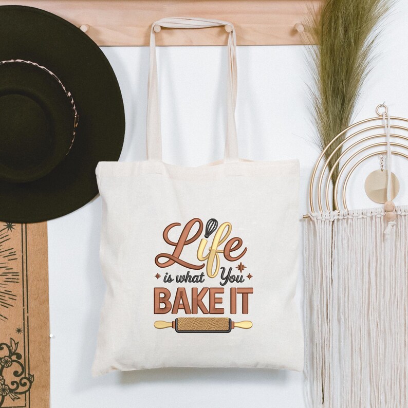Pode incluir: Saco tote creme com o texto "Life is what you bake it" em letras castanhas e amarelas. O design inclui um batedor e um rolo de massa. O saco tem al&ccedil;as creme e est&aacute; pendurado num cabide de madeira.
