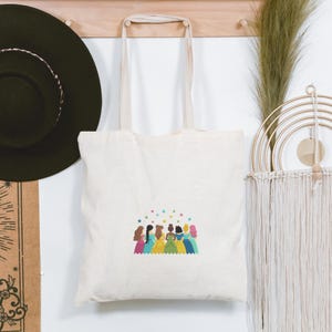 May include: Cream-colored tote bag featuring a colorful illustration of several figures in gowns, with stars above them. The bag has long, cream-colored handles and is hanging on a wooden rack. A black hat and decorative wall hangings are also visible.