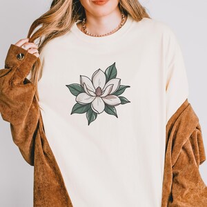 Pode incluir: Camisola creme com um design floral detalhado. O gr&aacute;fico mostra uma flor de magn&oacute;lia branca com folhas verdes e um centro rosa. A camisola &eacute; combinada com um casaco castanho.