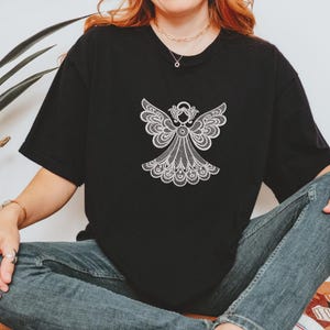 May include: Black t-shirt featuring a white, detailed angel design. The angel has outstretched wings and a decorative halo. The design is centered on the front of the shirt. The shirt is a casual style.