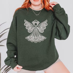 May include: A dark green sweatshirt featuring a detailed white angel design on the front. The angel has intricate wing and dress details, creating a delicate, lace-like appearance. The sweatshirt is a solid color, offering a simple backdrop for the detailed graphic.